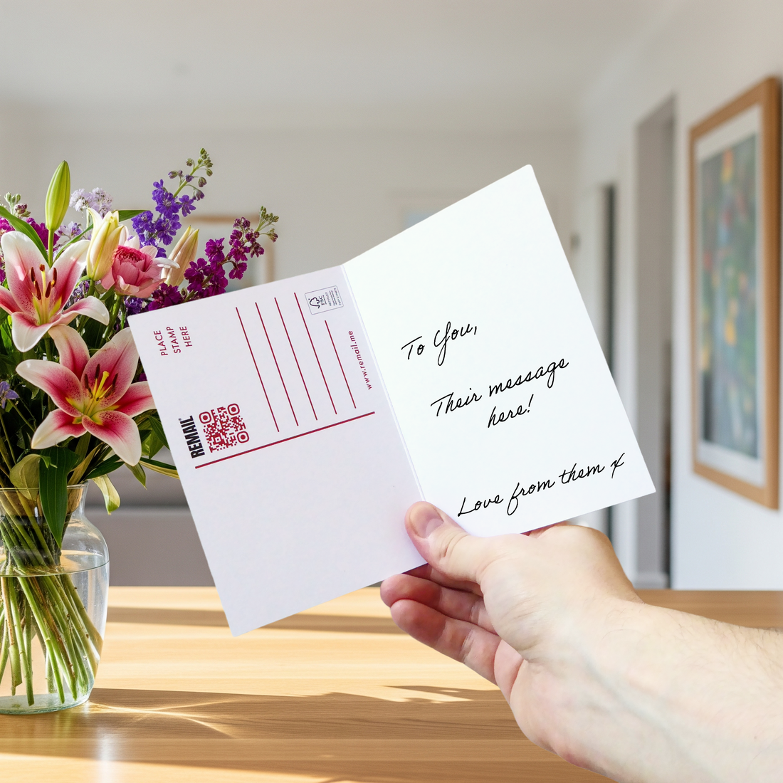 Open reusable greeting card held in one hand, showing a handwritten message on the right and a detachable postcard layout on the left, beside a vase of flowers.